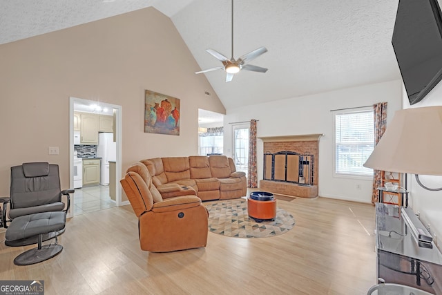 living room with a ceiling fan, light wood-style flooring, a textured ceiling, a fireplace, and high vaulted ceiling