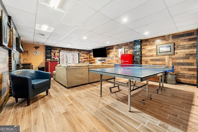 game room featuring light wood finished floors, a drop ceiling, and wood walls