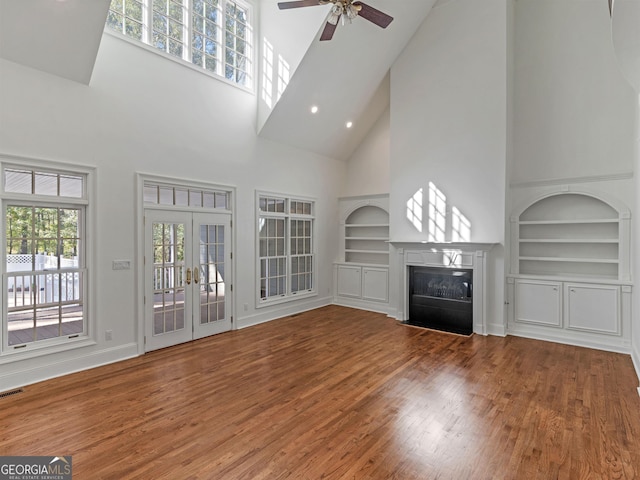 unfurnished living room featuring ceiling fan, wood finished floors, a fireplace with flush hearth, built in features, and french doors