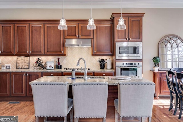 kitchen with appliances with stainless steel finishes, ornamental molding, light stone countertops, light wood-type flooring, and under cabinet range hood