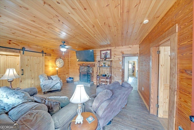 living area featuring a barn door, wooden walls, a ceiling fan, wood ceiling, and a stone fireplace