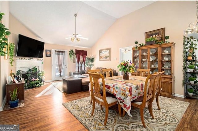 dining area with ceiling fan with notable chandelier, a fireplace, wood finished floors, baseboards, and vaulted ceiling