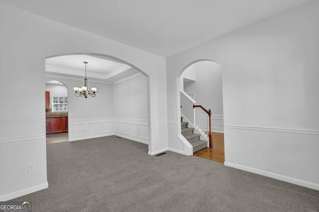 unfurnished room with a raised ceiling, a chandelier, a wainscoted wall, stairway, and dark carpet