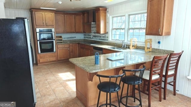 kitchen featuring a peninsula, a sink, wall chimney range hood, appliances with stainless steel finishes, and brown cabinetry