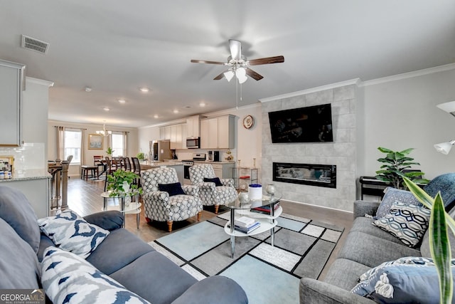 living room featuring visible vents, light wood-style flooring, a fireplace, ornamental molding, and ceiling fan with notable chandelier