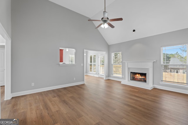 unfurnished living room with a ceiling fan, wood finished floors, high vaulted ceiling, a warm lit fireplace, and baseboards