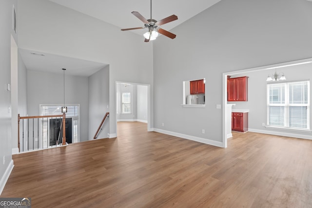 unfurnished living room featuring ceiling fan with notable chandelier, light wood-type flooring, a towering ceiling, and baseboards