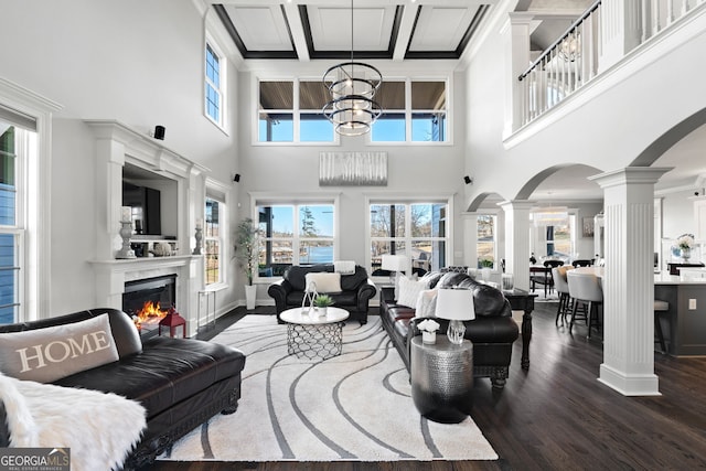 living room featuring arched walkways, dark wood-type flooring, a high end fireplace, ornate columns, and an inviting chandelier