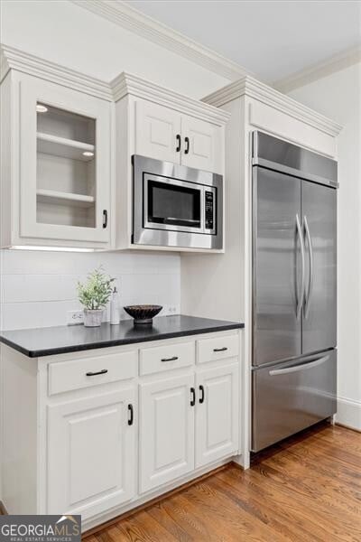 kitchen featuring built in appliances, light wood-style flooring, white cabinetry, dark countertops, and crown molding