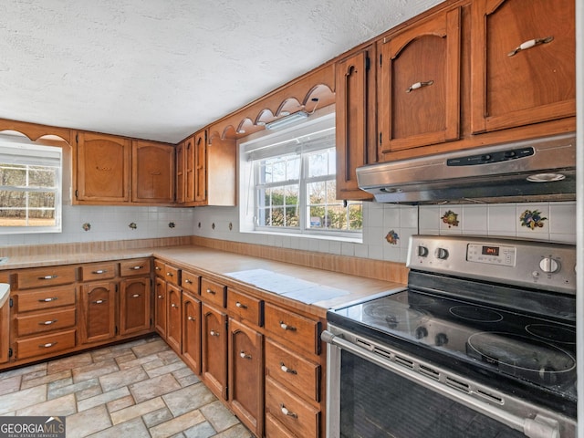 kitchen featuring stainless steel electric stove, light countertops, brown cabinetry, a healthy amount of sunlight, and under cabinet range hood