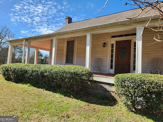 doorway to property with a shingled roof, covered porch, a yard, and a chimney