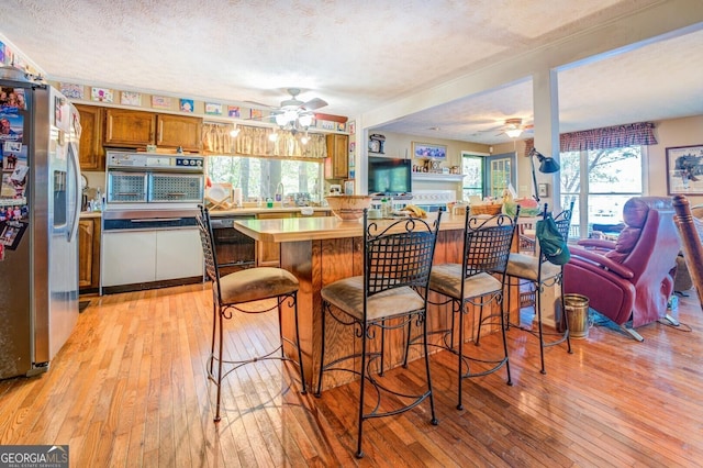 kitchen with wall oven, light wood-style flooring, open floor plan, stainless steel refrigerator with ice dispenser, and brown cabinetry