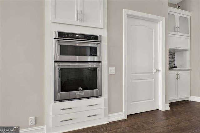 kitchen featuring double oven, dark wood-type flooring, glass insert cabinets, white cabinetry, and baseboards