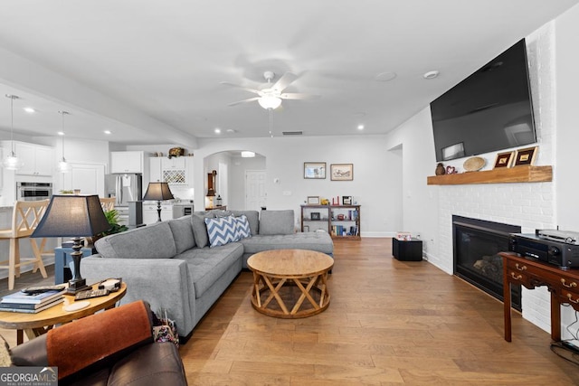 living area with recessed lighting, arched walkways, light wood finished floors, a brick fireplace, and ceiling fan