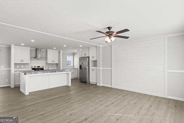 kitchen featuring white cabinets, a ceiling fan, wall chimney exhaust hood, appliances with stainless steel finishes, and light wood-type flooring