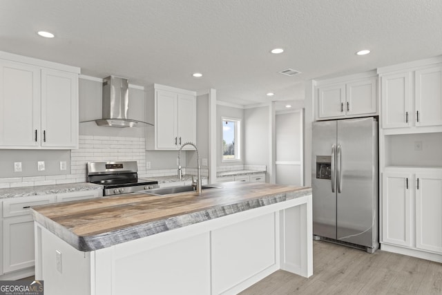 kitchen with stainless steel appliances, a sink, wood counters, wall chimney range hood, and an island with sink