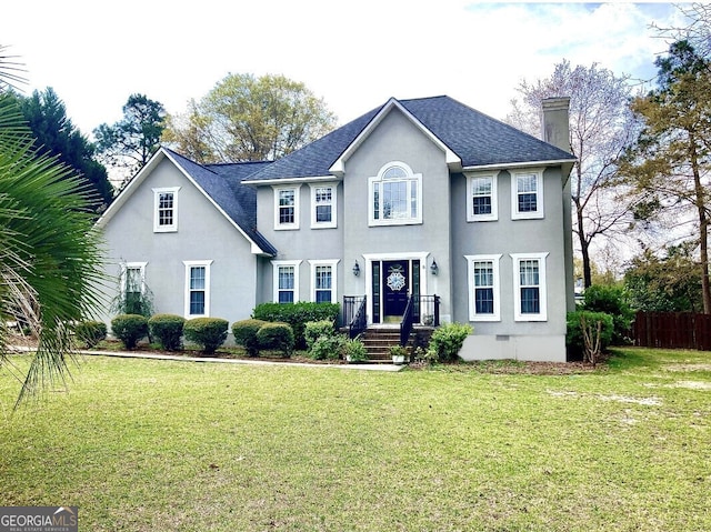 colonial house with a shingled roof, a front yard, a chimney, and stucco siding