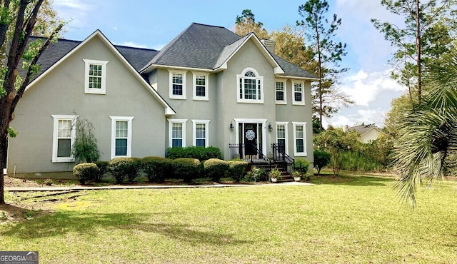 colonial-style house with a front yard, roof with shingles, a chimney, and stucco siding