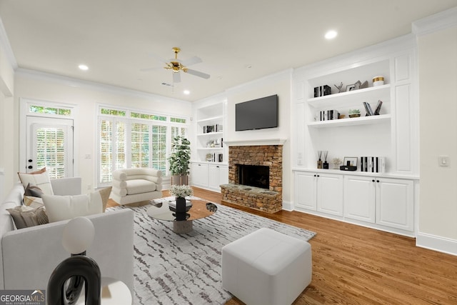 living area featuring ceiling fan, a stone fireplace, built in shelves, ornamental molding, and light wood finished floors