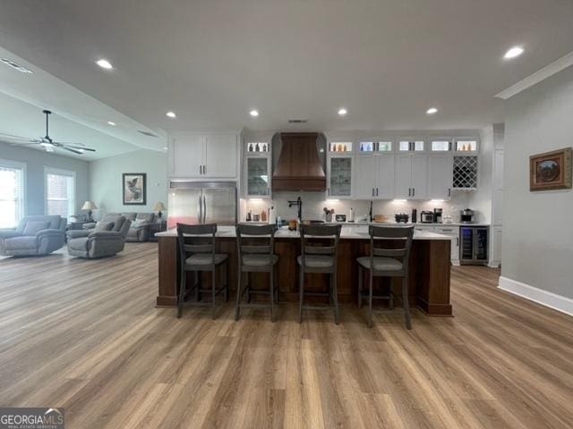 kitchen featuring beverage cooler, premium range hood, built in refrigerator, white cabinets, and a large island with sink