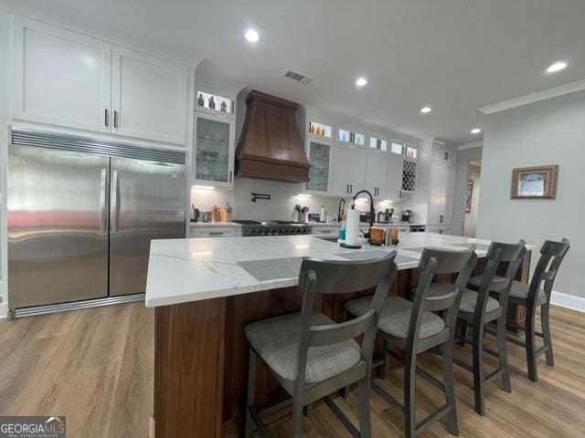 kitchen featuring visible vents, light wood-style floors, stainless steel built in fridge, an island with sink, and custom range hood