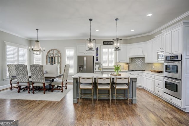 kitchen featuring stainless steel appliances, light countertops, custom range hood, a kitchen island with sink, and wood finished floors