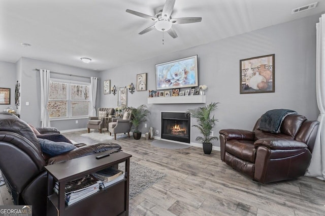 living area featuring baseboards, visible vents, a fireplace with flush hearth, ceiling fan, and wood finished floors