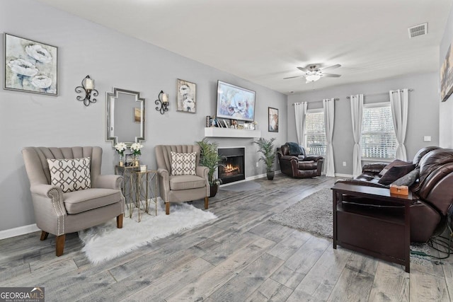 living room with a warm lit fireplace, baseboards, visible vents, ceiling fan, and wood finished floors