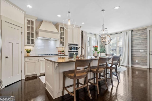 kitchen featuring premium range hood, appliances with stainless steel finishes, dark wood finished floors, and a center island with sink