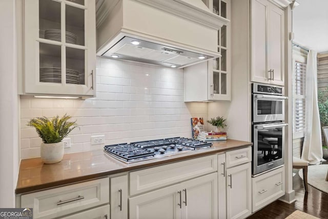 kitchen with glass insert cabinets, white cabinetry, appliances with stainless steel finishes, and custom exhaust hood