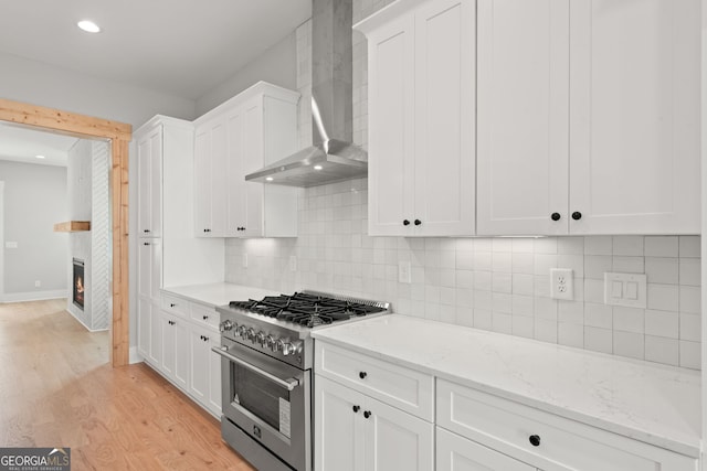 kitchen featuring high end stove, a fireplace, white cabinets, light wood-style floors, and wall chimney range hood