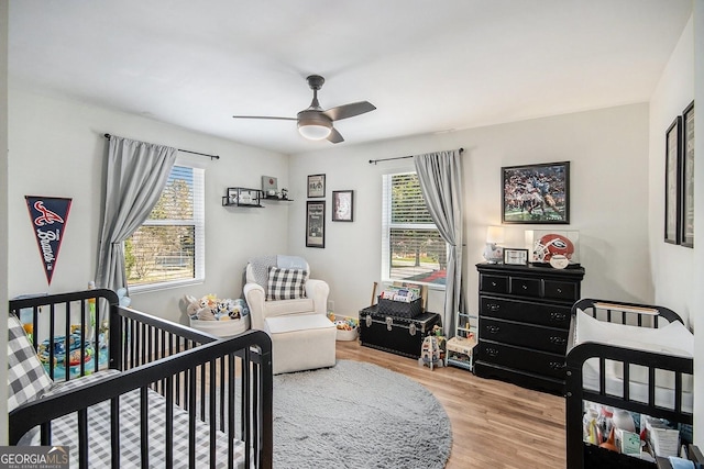 bedroom featuring a nursery area, ceiling fan, multiple windows, and wood finished floors