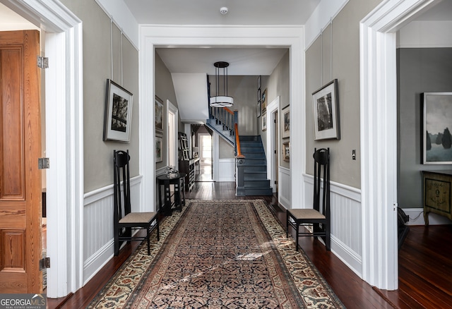 corridor with dark wood-type flooring, stairway, wainscoting, and a decorative wall