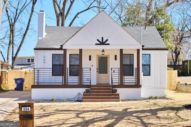 bungalow-style house featuring a shingled roof, a chimney, covered porch, fence, and board and batten siding