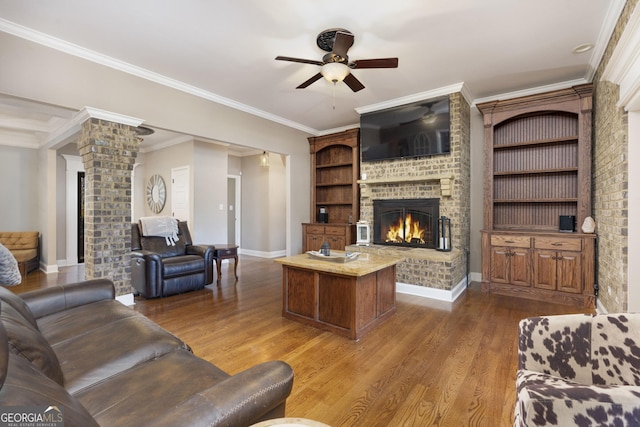 living area featuring wood finished floors, a ceiling fan, baseboards, a brick fireplace, and decorative columns