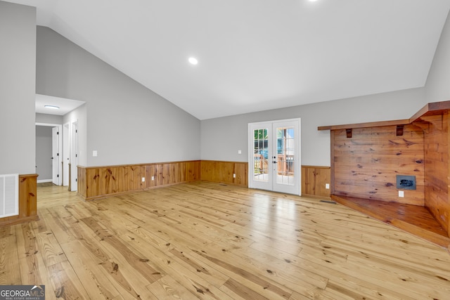 unfurnished living room with french doors, a wainscoted wall, light wood-style flooring, vaulted ceiling, and wooden walls