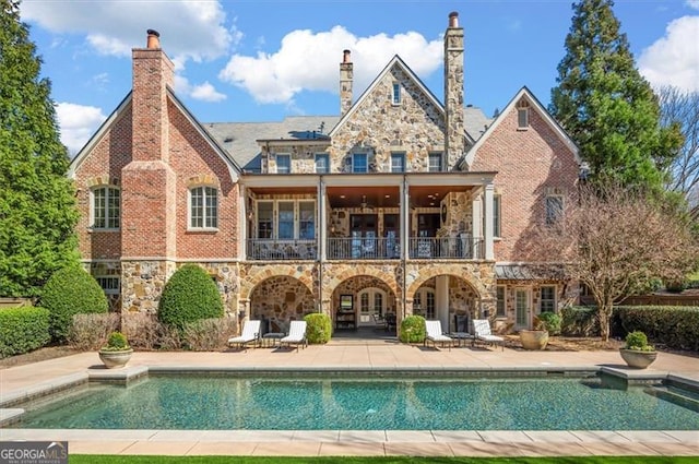 rear view of property featuring stone siding, a balcony, a patio area, an outdoor pool, and french doors
