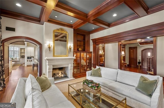 living area with arched walkways, beam ceiling, a fireplace with flush hearth, coffered ceiling, and wood finished floors
