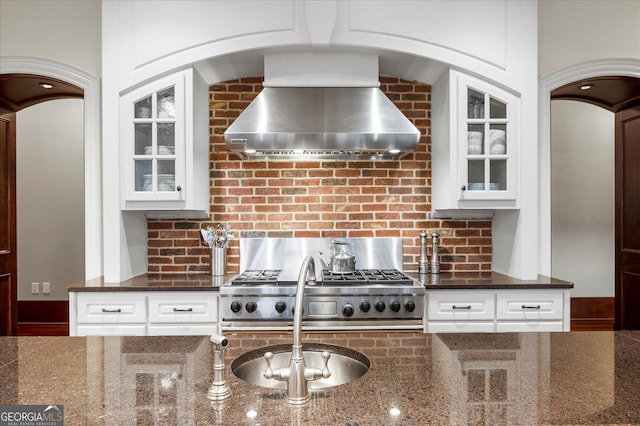 kitchen featuring dark stone counters, wall chimney range hood, glass insert cabinets, and decorative backsplash