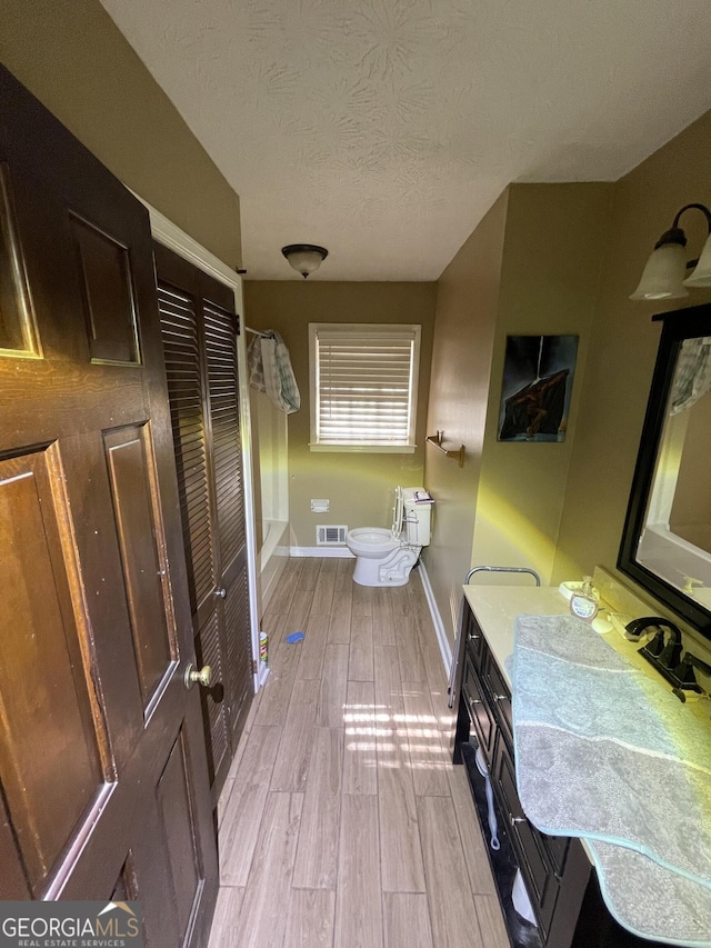 bathroom with a textured ceiling, toilet, wood finished floors, visible vents, and baseboards