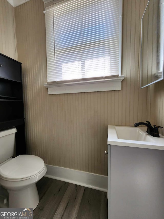 half bath featuring wooden walls, baseboards, toilet, wood finished floors, and vanity