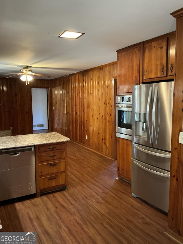 kitchen featuring brown cabinets, appliances with stainless steel finishes, dark wood-type flooring, a ceiling fan, and wooden walls