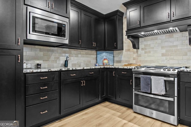 kitchen featuring light stone countertops, stainless steel appliances, dark cabinetry, light wood-type flooring, and decorative backsplash