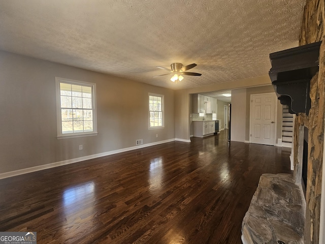 unfurnished living room featuring ceiling fan, a textured ceiling, a stone fireplace, dark wood-type flooring, and baseboards