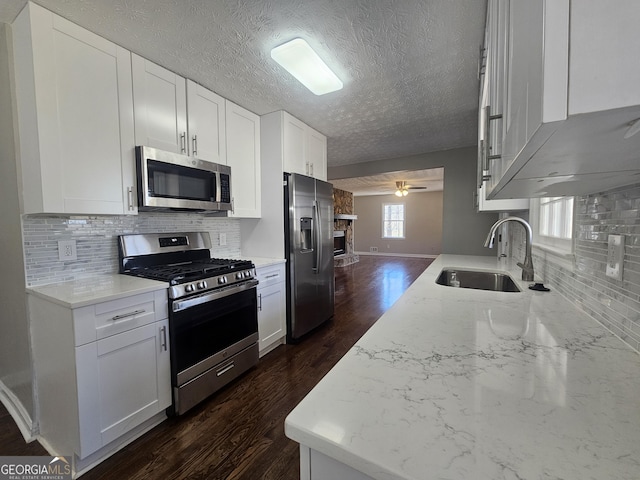 kitchen featuring a stone fireplace, stainless steel appliances, dark wood-type flooring, a sink, and a ceiling fan