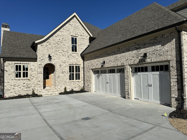 french country inspired facade featuring concrete driveway, an attached garage, brick siding, and roof with shingles