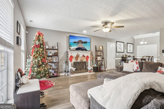 living room with visible vents, a fireplace with raised hearth, wood finished floors, a textured ceiling, and ceiling fan with notable chandelier