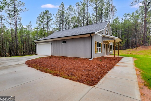 view of property exterior featuring roof with shingles, driveway, a view of trees, and an attached garage