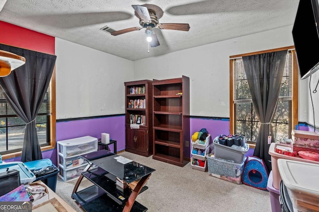 carpeted living area featuring a ceiling fan, a wainscoted wall, visible vents, and a textured ceiling