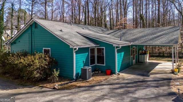 view of property exterior featuring central AC unit, an attached carport, a shingled roof, fence, and driveway